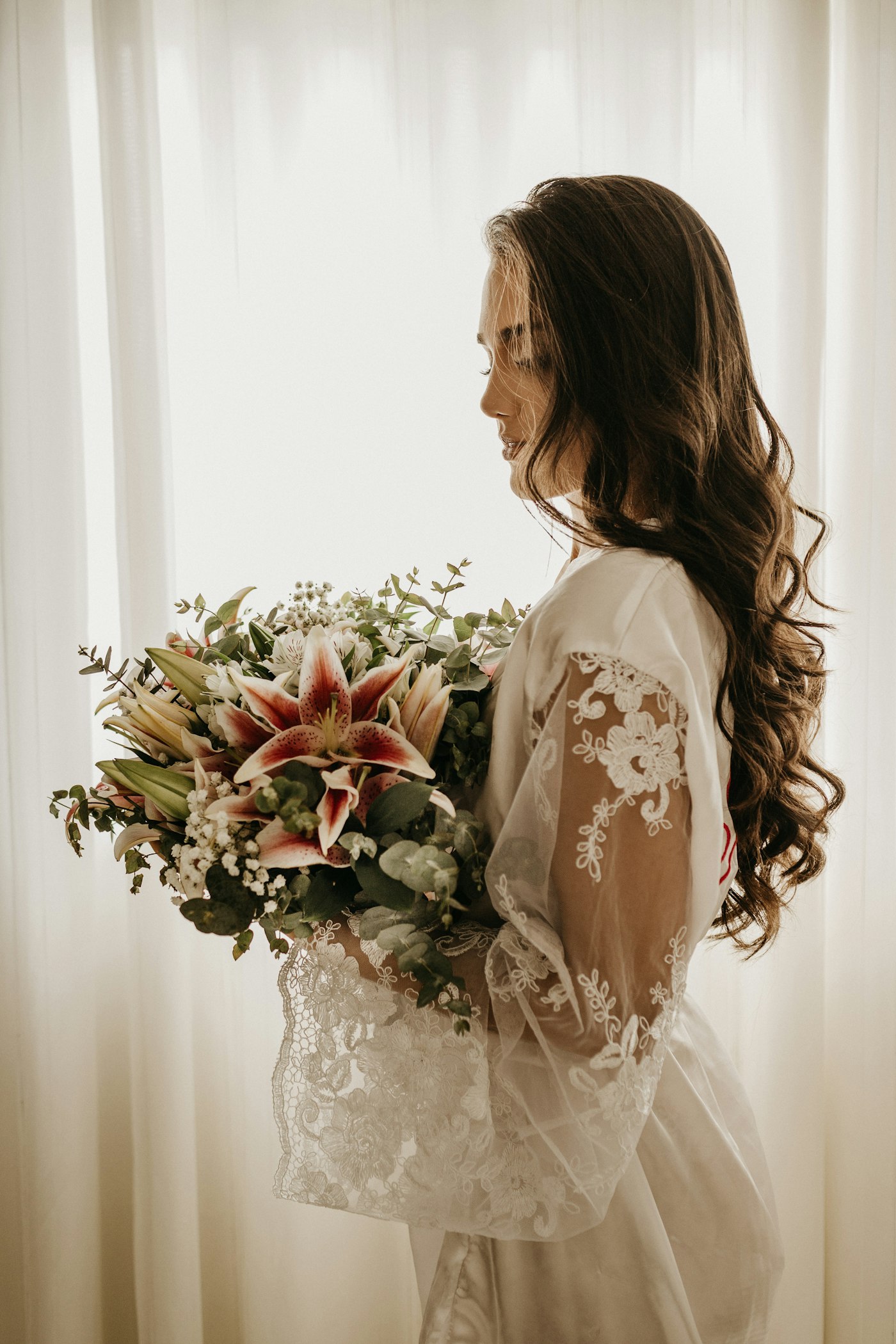 Mariée aux cheveux longs ondulés, tenant un bouquet près d’une fenêtre